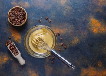 A imagem apresenta grãos de café, um recipiente de vidro e um fouet.