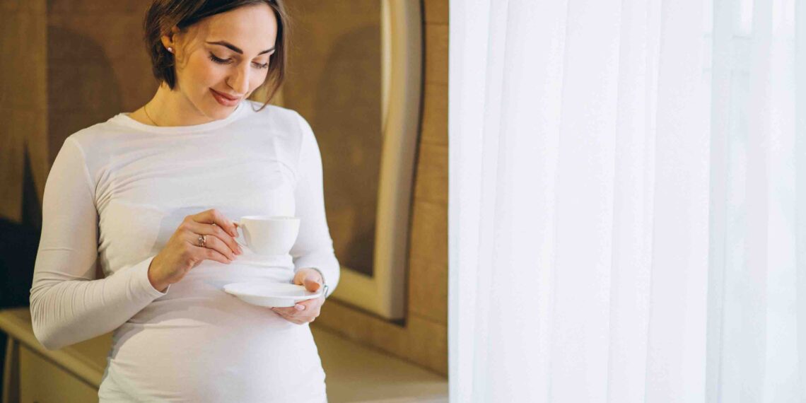 A imagem mostra uma mulher grávida com uma xícara na mão.